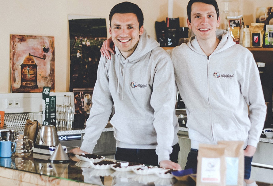Die beiden Gründer von earlybird coffee Emanuel und Merlin stehen hinter einer Kaffeebar, auf der frische Kaffeebohnen und earlybird Kaffee steht.