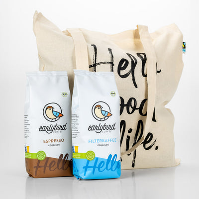 Eine Packung earlybird Filterkaffee gemahlen und eine Packung earlybird Espresso gemahlen stehen auf dem earlybird Jutebeutel, auf dem der Slogan "Hello good life" abgebildet ist
